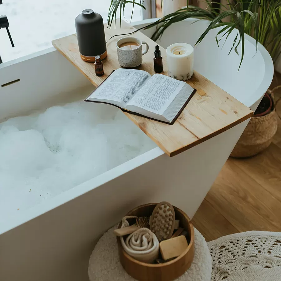 A relaxing bath scene with a white tub filled with bubbles. A wooden tray holds an open book, a mug, candles, and bottles. Nearby is a basket with towels and bath accessories; green plants decorate the space.