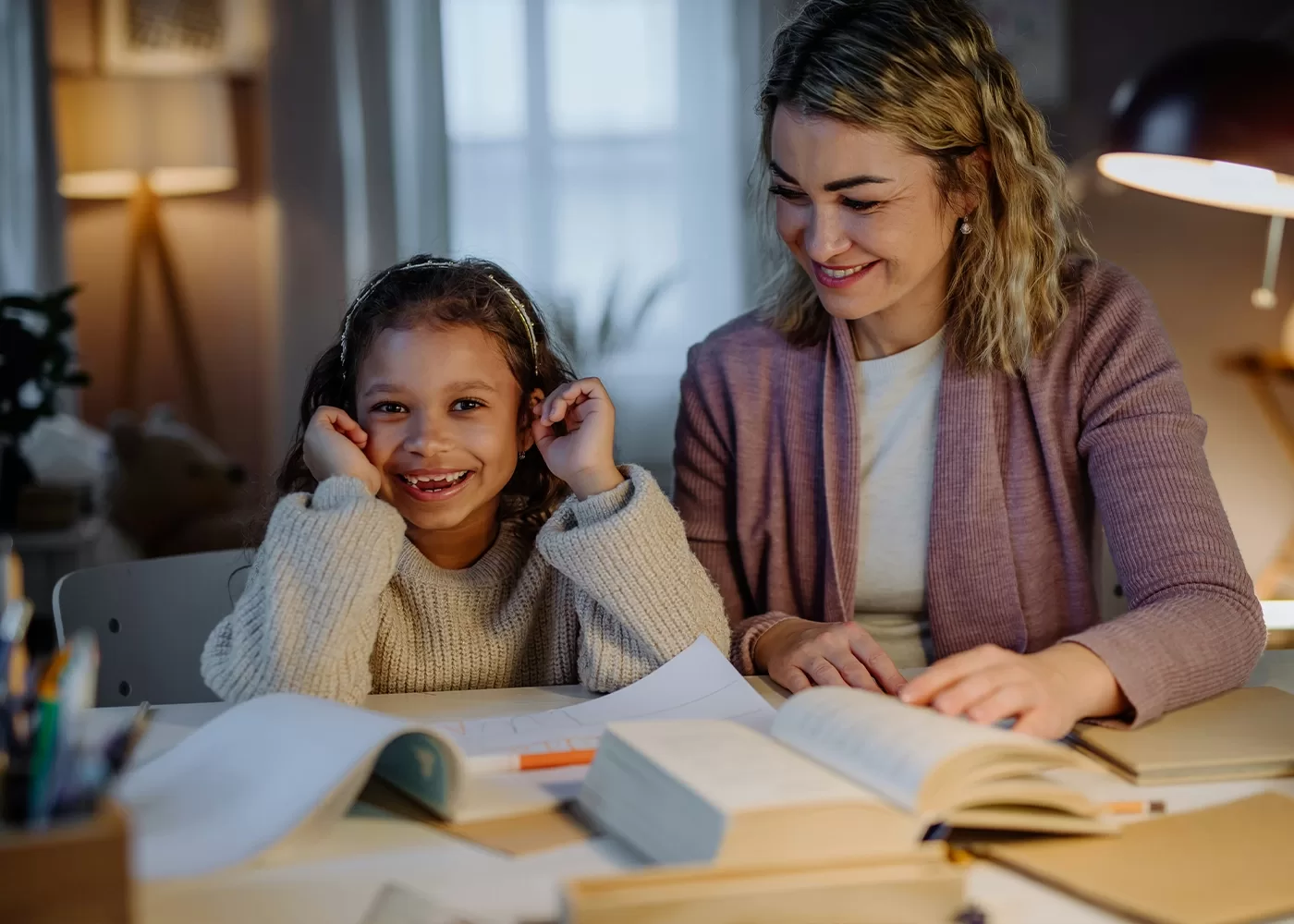 A smiling young girl sits at a table with an open notebook, while an adult woman beside her reads a book and smiles. The scene appears warm and cozy, with books and a lamp in the background.