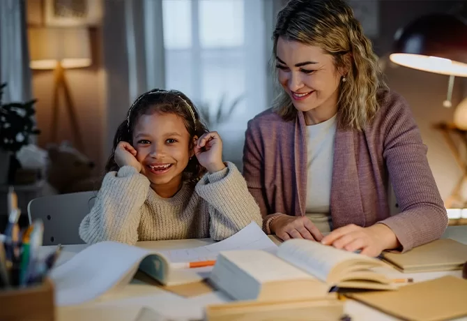 A smiling young girl sits at a table with an open book, resting her hands on her cheeks, while a woman beside her, also smiling, helps with homework. The scene is warmly lit, suggesting a cozy evening study session.
