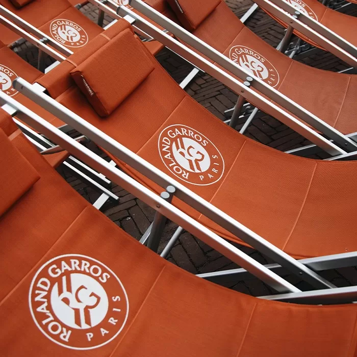 Several orange lounge chairs with the Roland Garros logo and Paris printed on the fabric are arranged closely together on a brick surface.