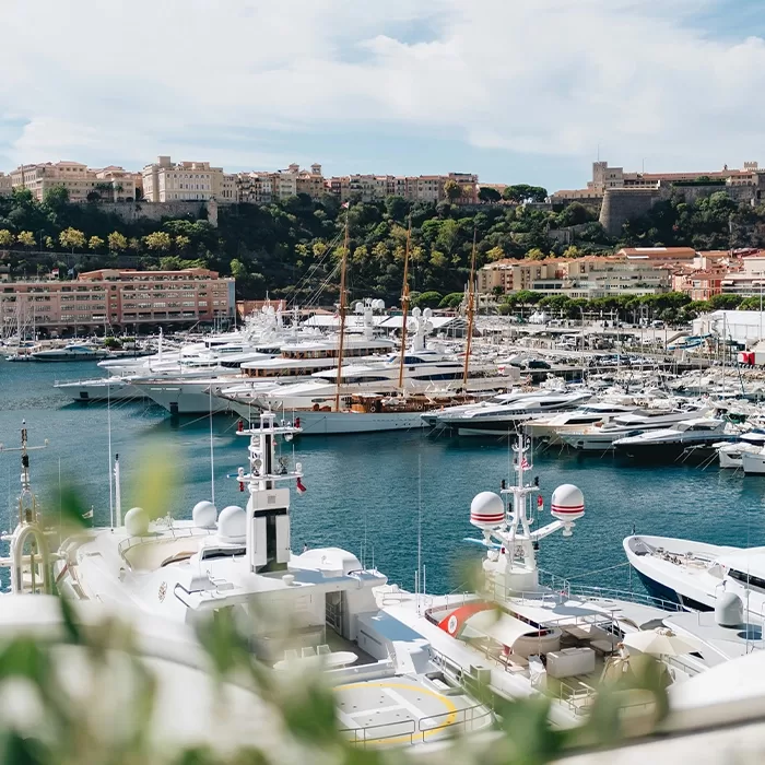 A scenic marina filled with luxury yachts docked in blue water, with a hillside town featuring historic buildings and lush greenery in the background under a partly cloudy sky.