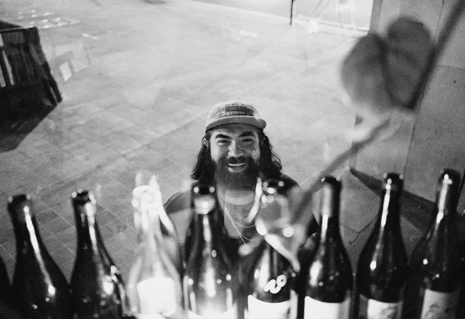A bearded man wearing a cap smiles while standing behind a row of assorted bottles on a counter, viewed through a window at night in a black and white photo.