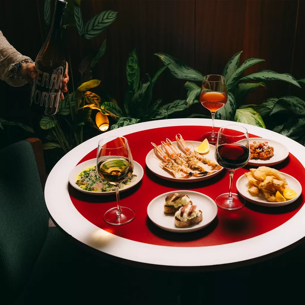 A round table with assorted tapas dishes, including prawns, chips, and bruschetta, plus three glasses of wine. A hand pours wine from a bottle. Lush green plants are in the background.
