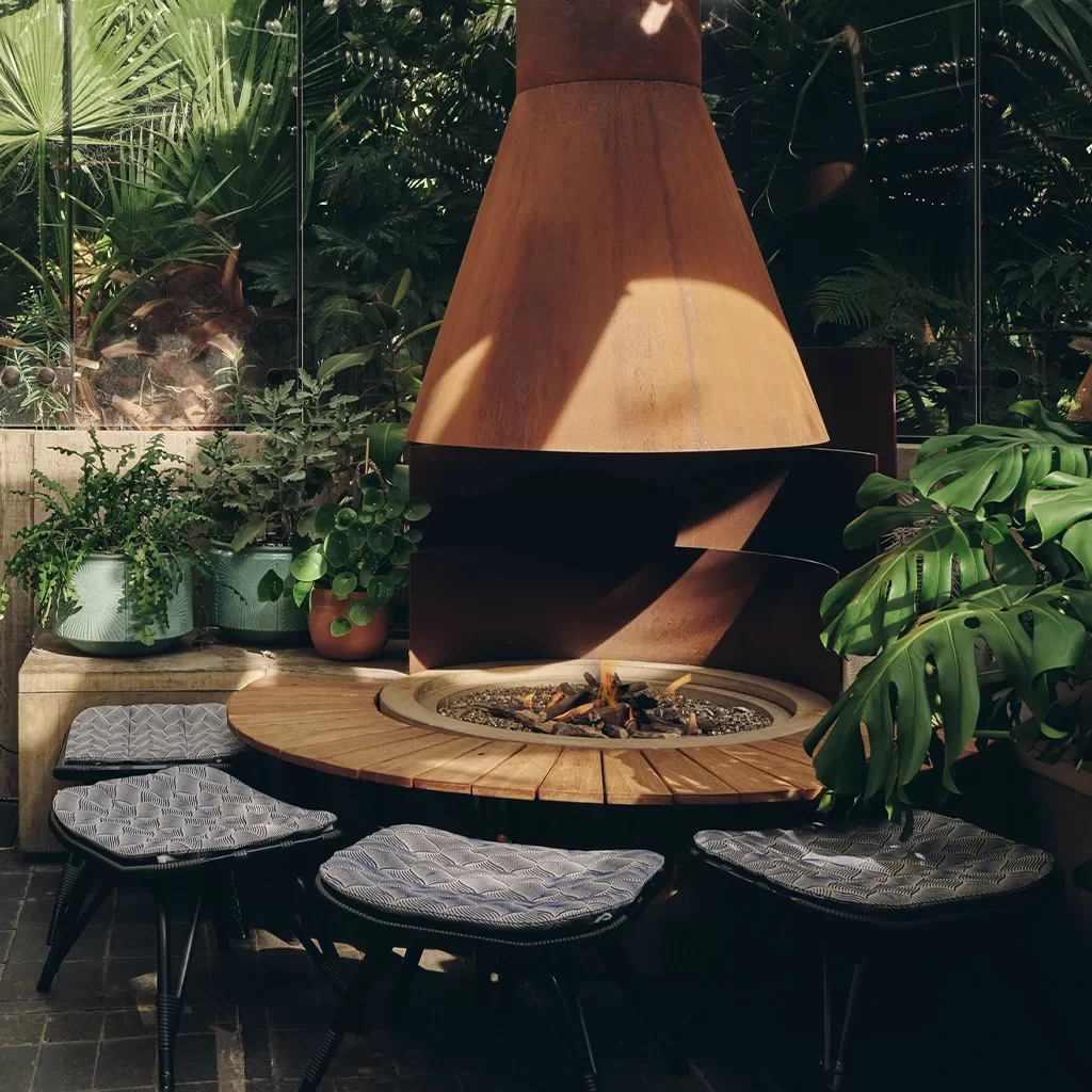 A cozy outdoor seating area with a round wooden table, modern stools with cushions, a central fire pit, potted plants, and a large rust-colored metal fireplace surrounded by lush greenery.
