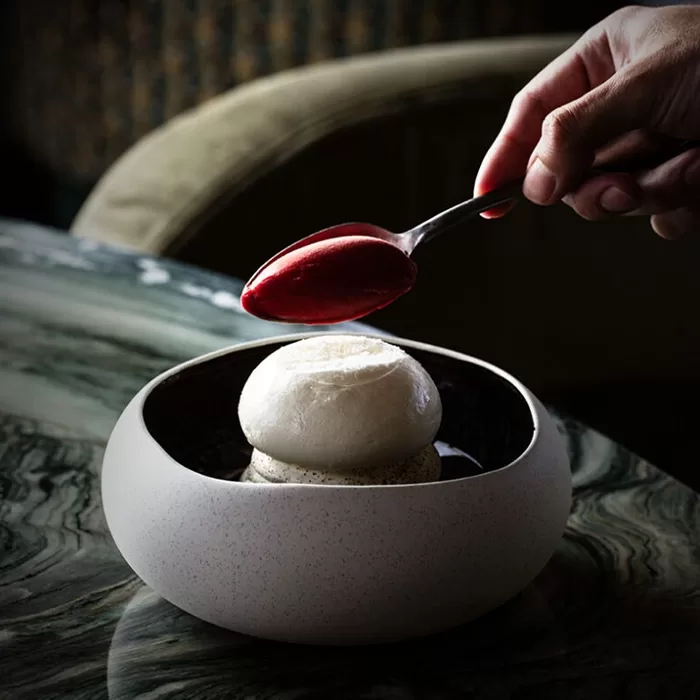A hand holds a spoonful of red sauce above a round white dessert served in a modern ceramic bowl on a marble table.