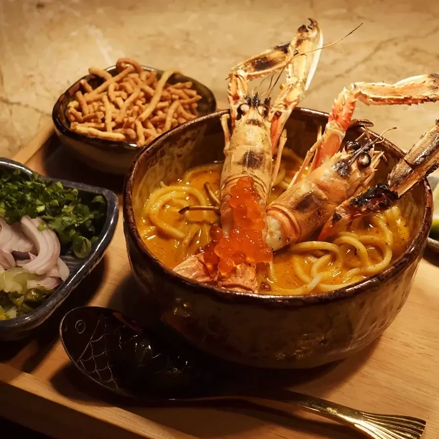 A bowl of creamy noodle soup topped with two large prawns and orange fish roe, served with sides of crispy noodles, sliced onions, green herbs, and a decorative spoon on a wooden tray.