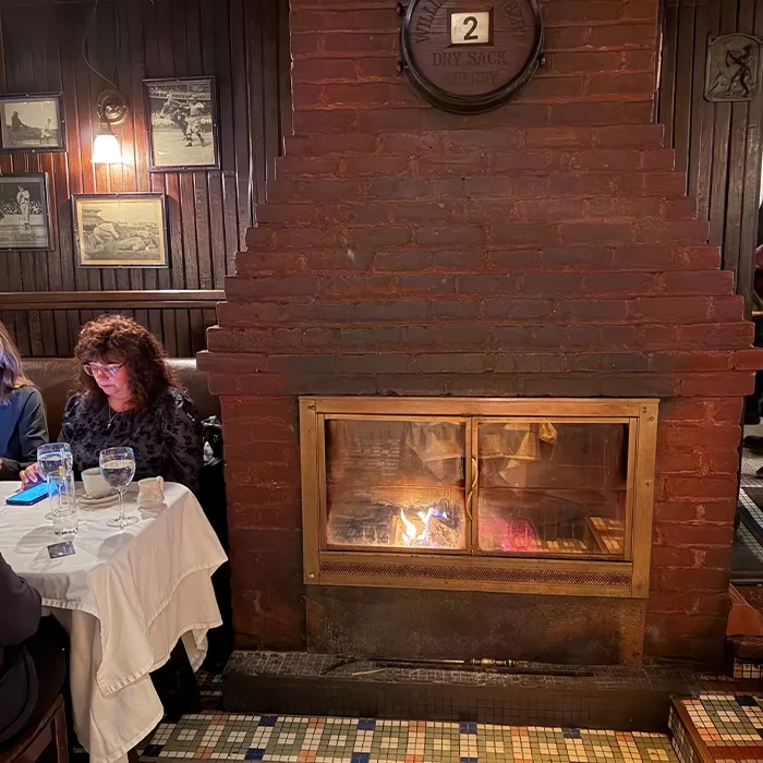A cozy restaurant scene with a brick fireplace in the center, a fire burning inside, tables set with white tablecloths and glasses, and people dining. The walls are adorned with framed photos and dark wood paneling.