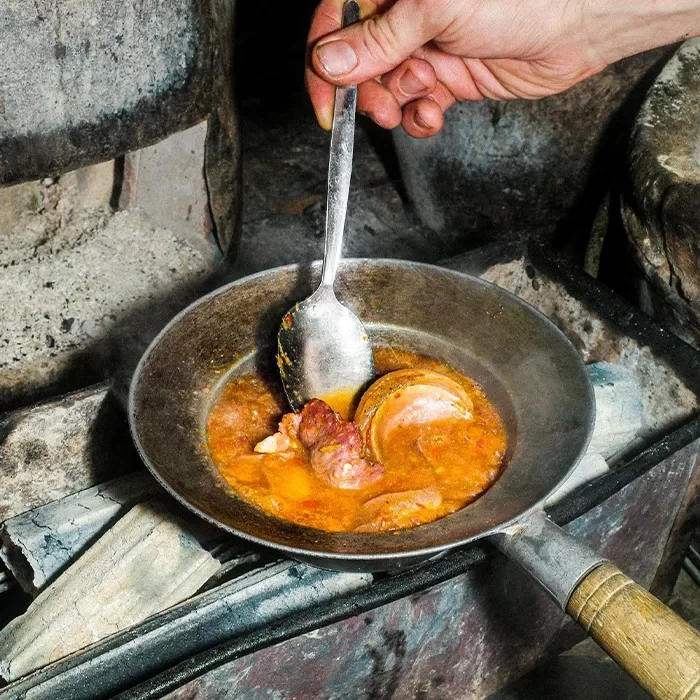 A hand holds a spoon over a rustic metal pan filled with a hearty stew, containing chunks of meat and vegetables, cooking on an old stove with wood beneath.