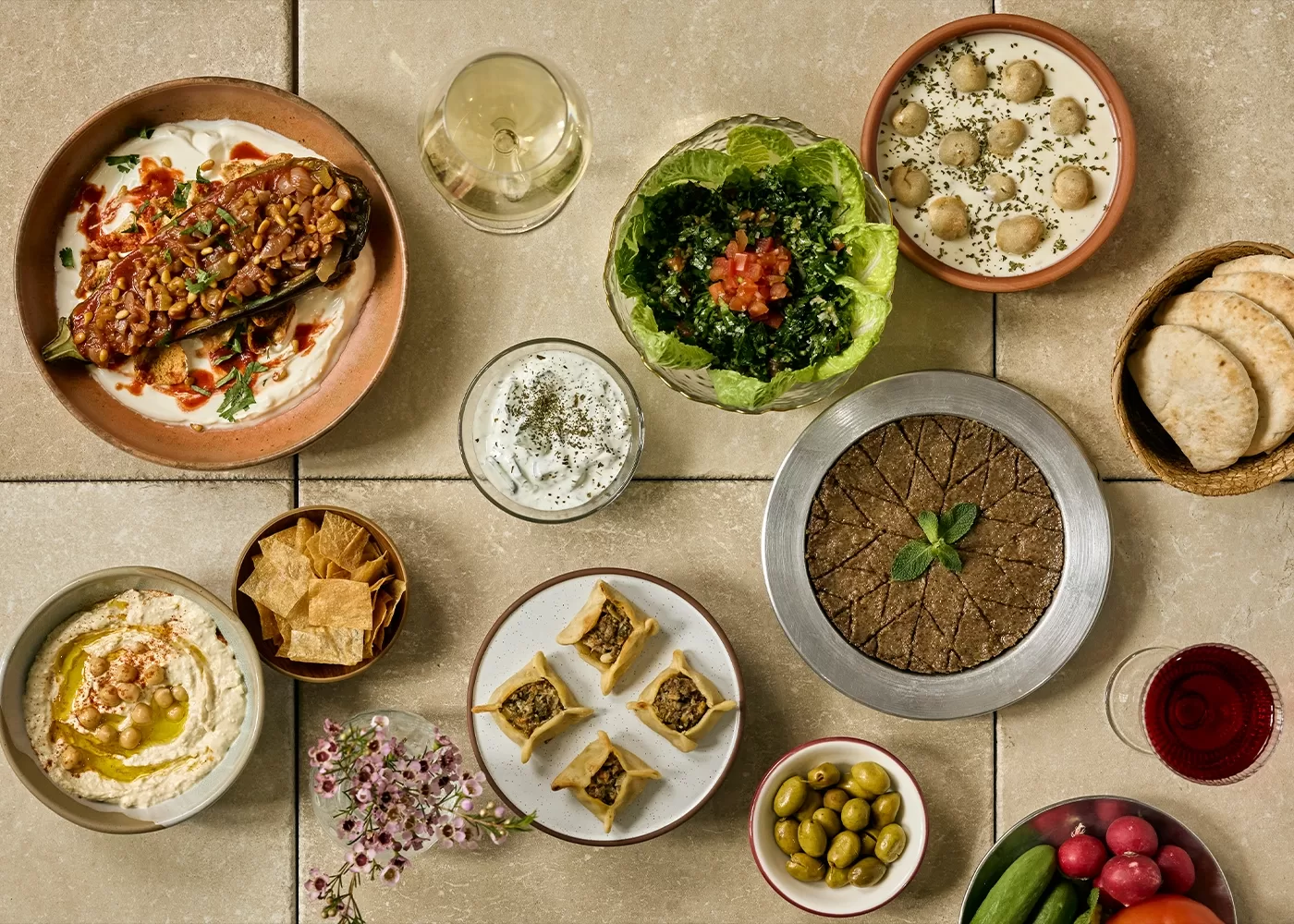 A table set with a variety of Middle Eastern dishes, including hummus, baba ganoush, kibbeh, stuffed eggplant, grape leaves, tabbouleh, pita bread, olives, dips, raw vegetables, and glasses of white and red wine.