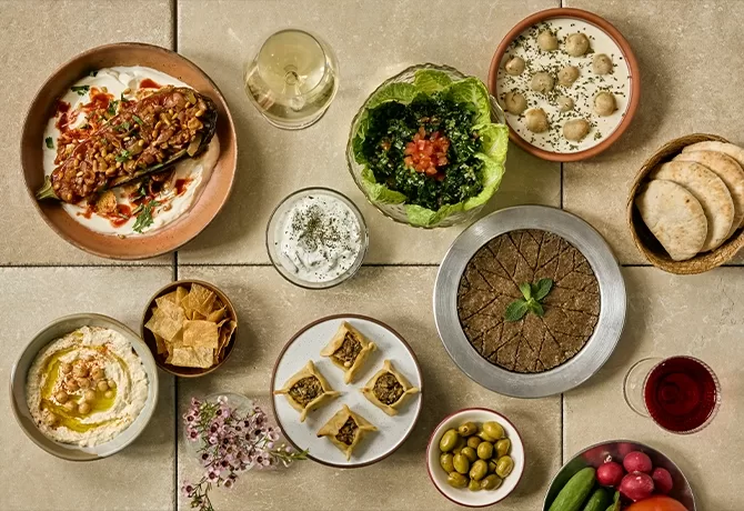 A variety of Middle Eastern dishes on a table, including hummus, kibbeh, baba ghanoush, salad in lettuce, pita bread, olives, yogurt dip, chopped vegetables, pastries, and glasses of wine.