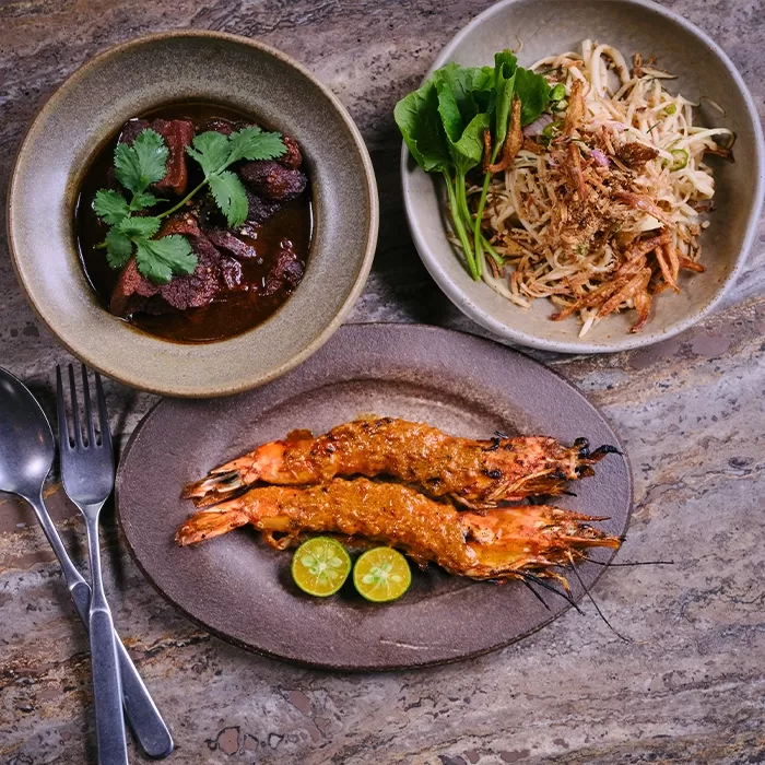 Three dishes on a stone table: two large grilled prawns with sauce and lime halves, a bowl of dark meat stew garnished with cilantro, and a bowl of salad with herbs and grated vegetables. Fork and spoon are beside the plates.