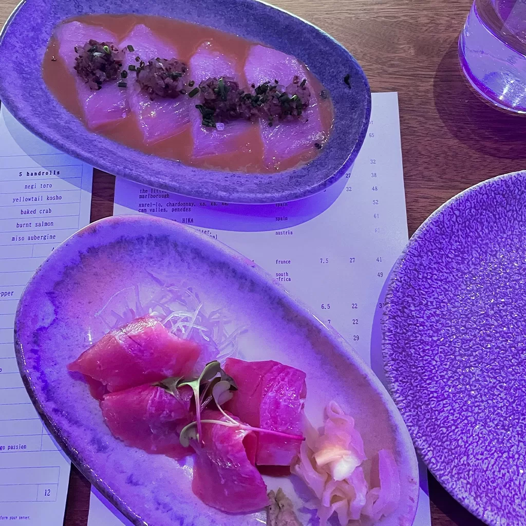 Two plates of sashimi are set on a table with a menu beneath them. One plate has slices topped with garnish in a sauce, and the other has plain pink slices, shredded daikon, pickled ginger, and wasabi. The lighting is purple.