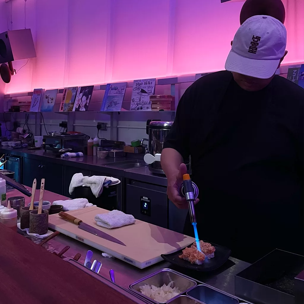 A chef in a white cap uses a blowtorch to sear sushi at a counter in a dimly lit restaurant with pink and purple ambient lighting and vinyl record covers displayed on the wall behind him.