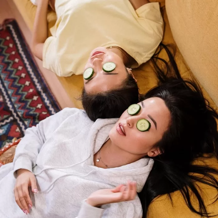 Two women are lying on a couch with cucumber slices on their eyes, relaxing. One wears a yellow shirt and the other a gray hoodie. They are on a yellow couch next to a patterned rug.