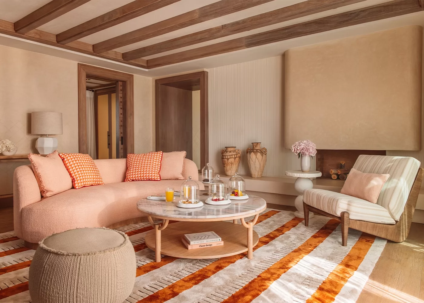 A cosy living room with a pink sofa and orange-checkered pillows. Theres a round coffee table with drinks and snacks, a striped armchair, and decorative vases. The room features wooden beams and a patterned rug in warm tones in Temuda Bay.
