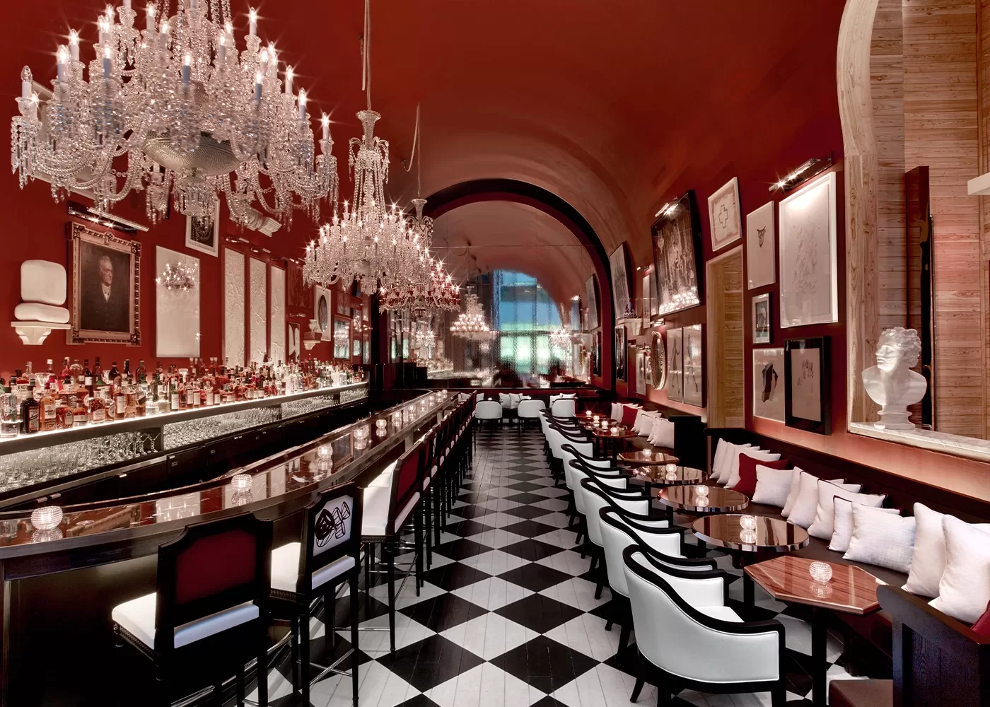 Luxurious restaurant interior with elegant chandeliers, a long bar on the left, and rows of tables with white chairs on the right. The room features a high ceiling and red walls adorned with framed artwork and mirrors. Black and white checkered floor tiles.