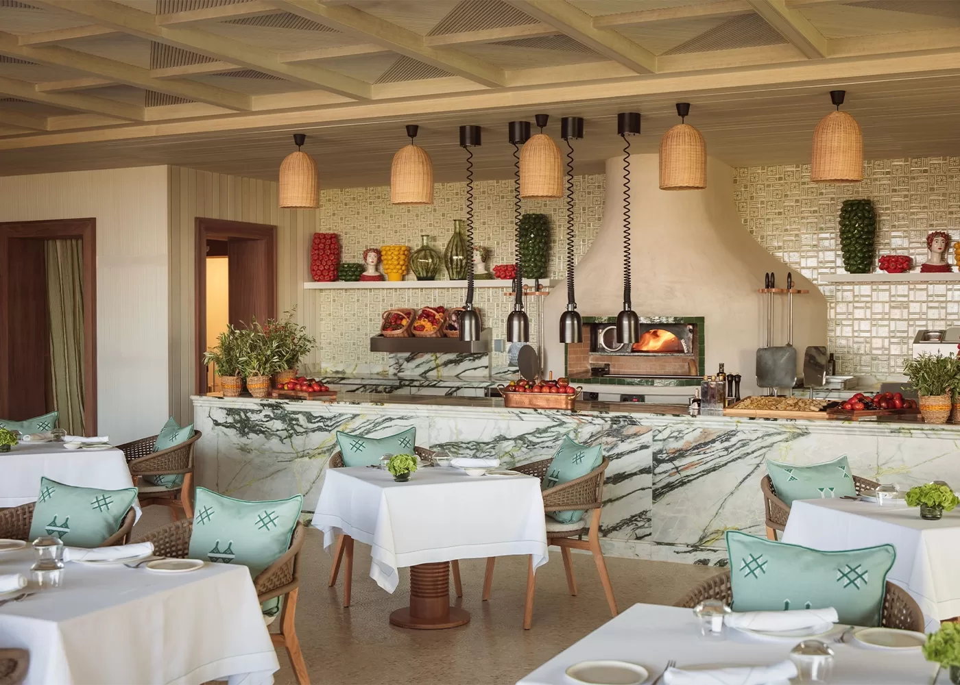 A cosy restaurant interior with white tablecloth-covered tables and turquoise cushions. A marble counter displays fresh produce, and an oven is in the background. Hanging wicker lamps add a warm touch, and the setting is elegant and inviting in in Temuda Bay.