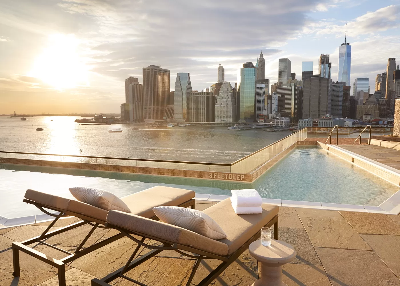 2 lounge chairs set out beside a pool overlooking the New York City skyline.
