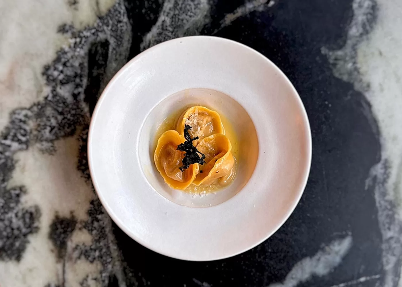 A white plate with three pieces of yellow ravioli, garnished with black shavings, sits on a dark, marbled tabletop.