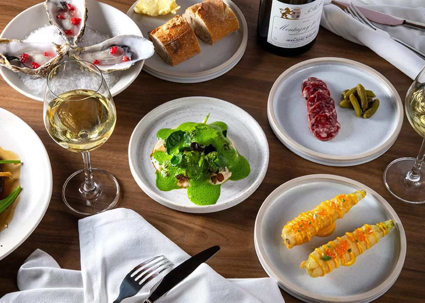 A table set with assorted dishes: oysters on ice, sliced baguette, a green-topped entrée, salami with pickles, corn with garnish, a glass of white wine, a wine bottle, and utensils on a white napkin.