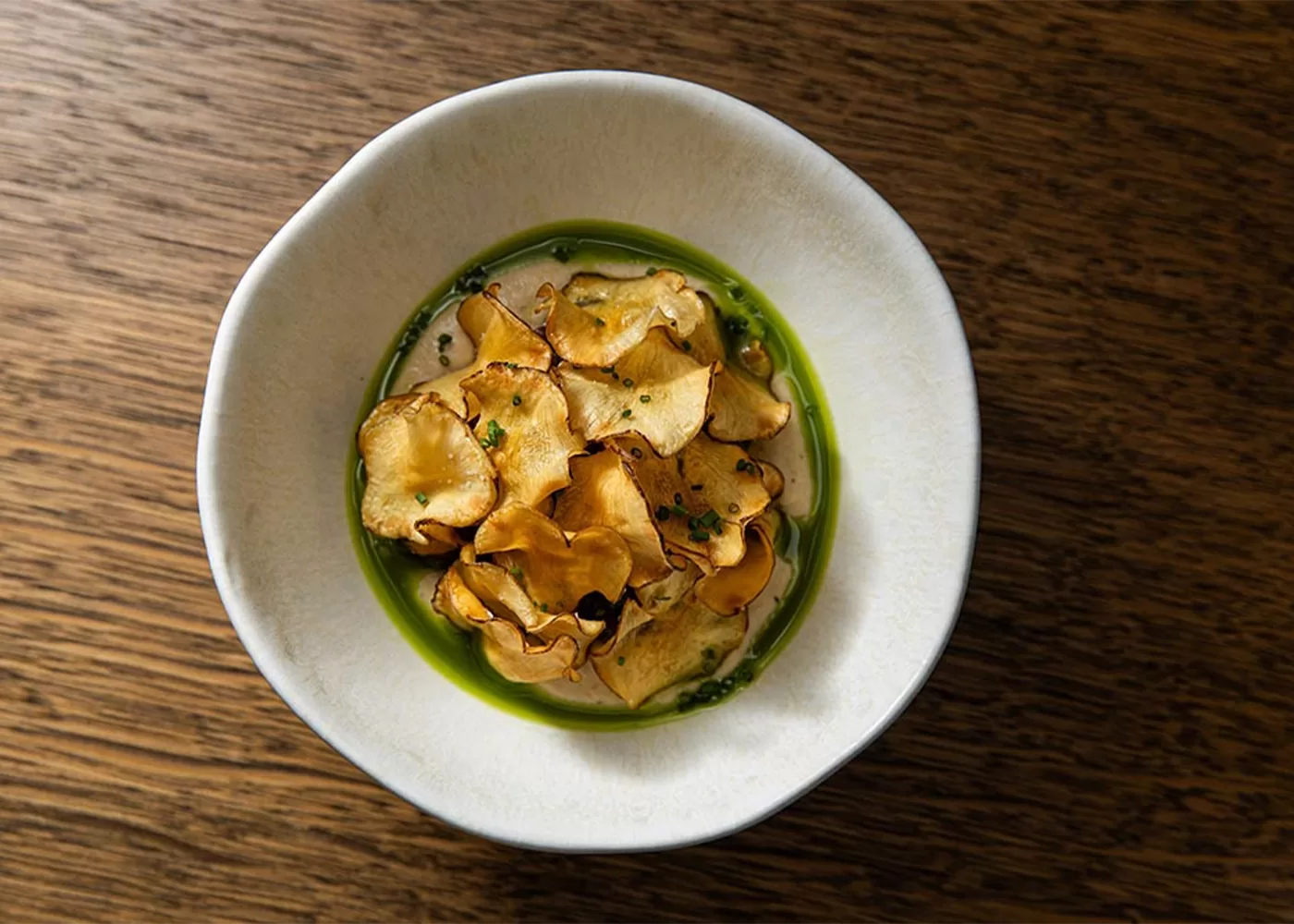 A white bowl filled with thin, golden-brown vegetable chips sits on a wooden surface. The chips are arranged over a green sauce, creating a visually appealing and appetizing presentation.