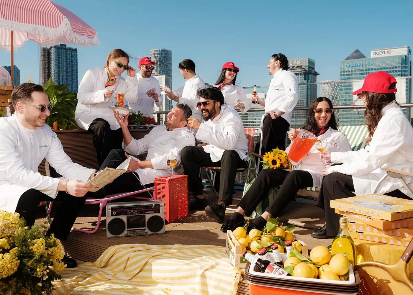 A group of people in white chef coats relax on a rooftop with drinks, snacks, and fruit baskets. Some are chatting or laughing, others read or listen to a boombox. City skyscrapers are visible in the background under a clear sky.