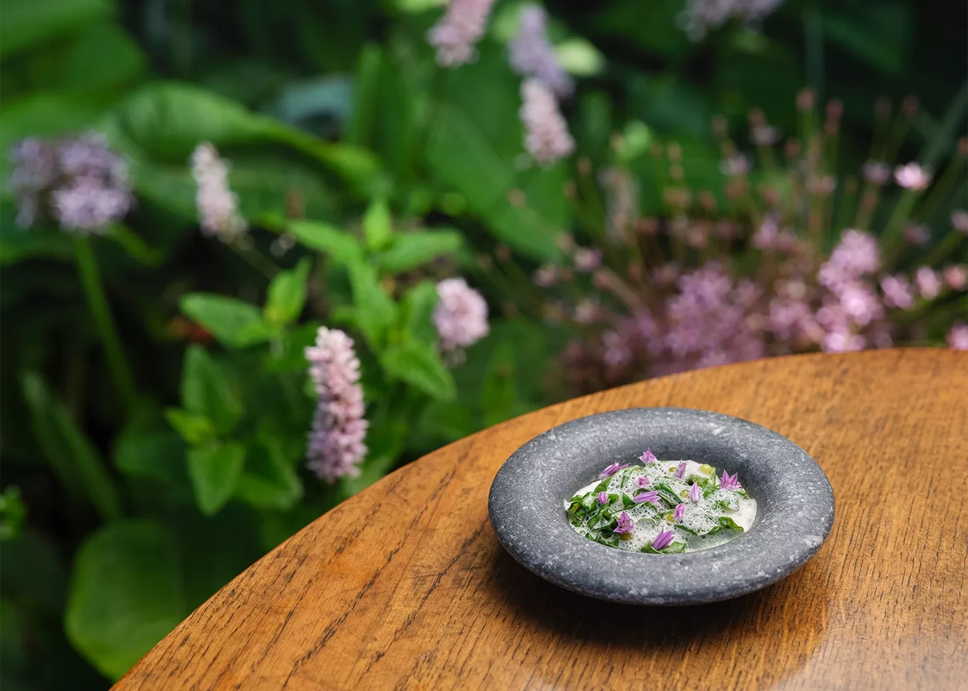 A gray stone plate with a small, colorful salad garnished with purple flowers sits on a wooden table, with lush green plants and pink flowers blurred in the background.
