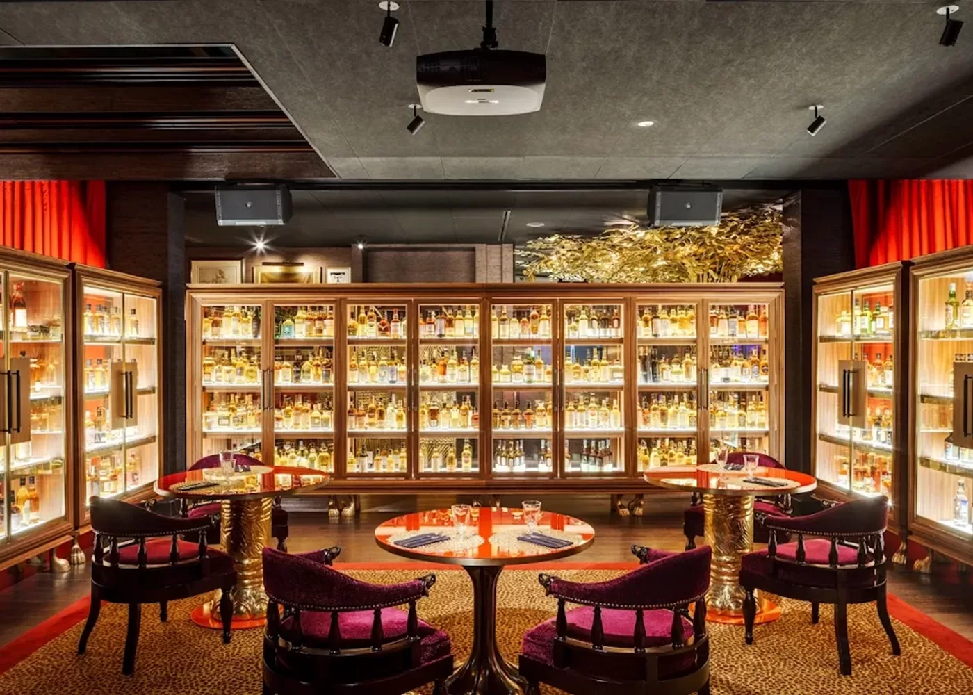 A stylish bar with shelves full of illuminated liquor bottles behind two round tables, each surrounded by plush, purple-cushioned chairs on a patterned carpet, creating a luxurious and inviting atmosphere.