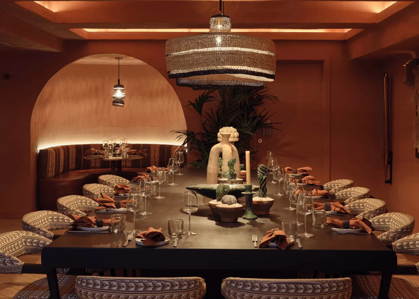A dimly lit, elegantly set dining room with a long table surrounded by patterned chairs. A large decorative light fixture hangs above, and the table is adorned with glassware, orange napkins, and intricate centerpieces. The walls feature warm brown tones at Ixchel.