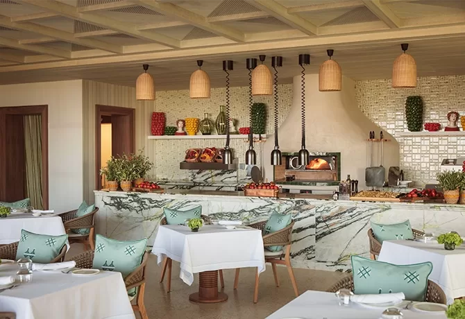 A cosy restaurant interior with white tablecloth-covered tables and turquoise cushions. A marble counter displays fresh produce, and an oven is in the background. Hanging wicker lamps add a warm touch, and the setting is elegant and inviting in in Temuda Bay.
