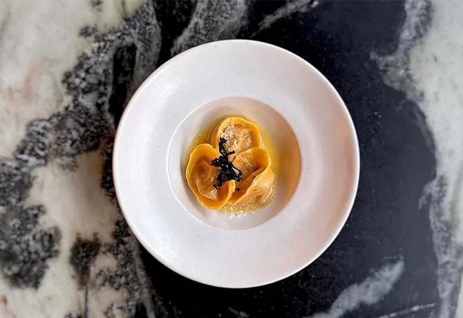 A white plate with three pieces of stuffed pasta topped with a small amount of black garnish, served in a light sauce on a marbled black and white table.