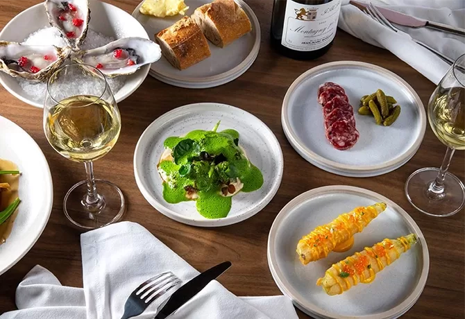 A table set with assorted gourmet dishes, including oysters, baguette slices, sausages with pickles, broccoli with green sauce, corn on the cob, two glasses of white wine, and a bottle of wine.