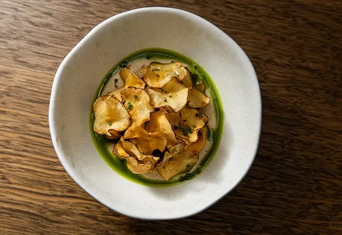 A white bowl filled with thin, crispy vegetable chips atop a green sauce, placed on a wooden surface.