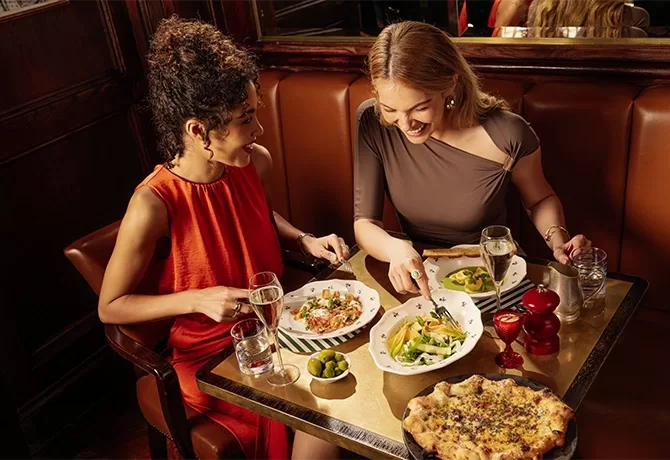 Two women sit at a restaurant table, smiling and enjoying a meal together. The table is set with plates of pasta, salad, bread, olives, glasses of water, and wine in an elegant, warmly lit setting.
