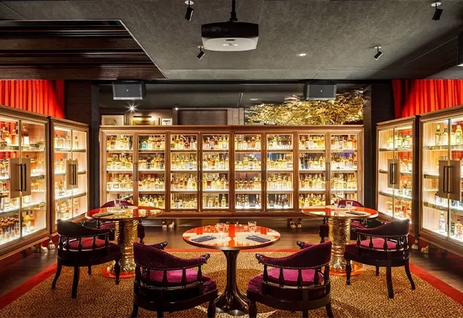 Luxurious bar lounge with four round tables, plush purple chairs, and walls lined with illuminated shelves displaying a vast collection of bottles. Warm lighting and elegant decor create a sophisticated atmosphere.