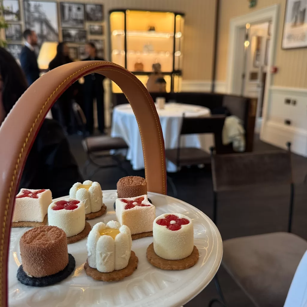 A plate of assorted mini desserts on a tiered stand in a stylish, softly lit café, with people and shelves visible in the background.