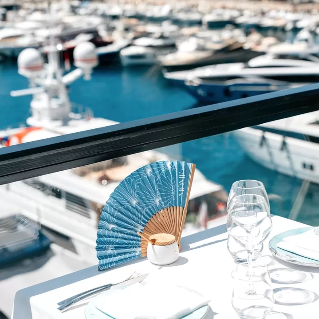 A table set with glasses, a blue decorative fan, and plates overlooks a marina filled with docked yachts and boats on a sunny day.