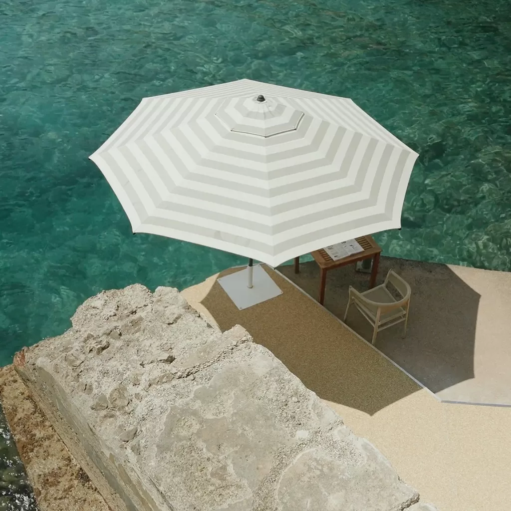A white and light gray striped umbrella shades a table and chair set on a stone patio beside clear turquoise water. Part of a stone wall is visible in the foreground.