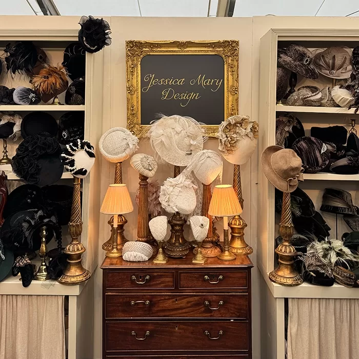 A vintage-style display features elegant hats on stands, surrounded by gold lamps and shelves filled with more hats. A framed sign above the setup reads Jessica Mary Design in cursive.