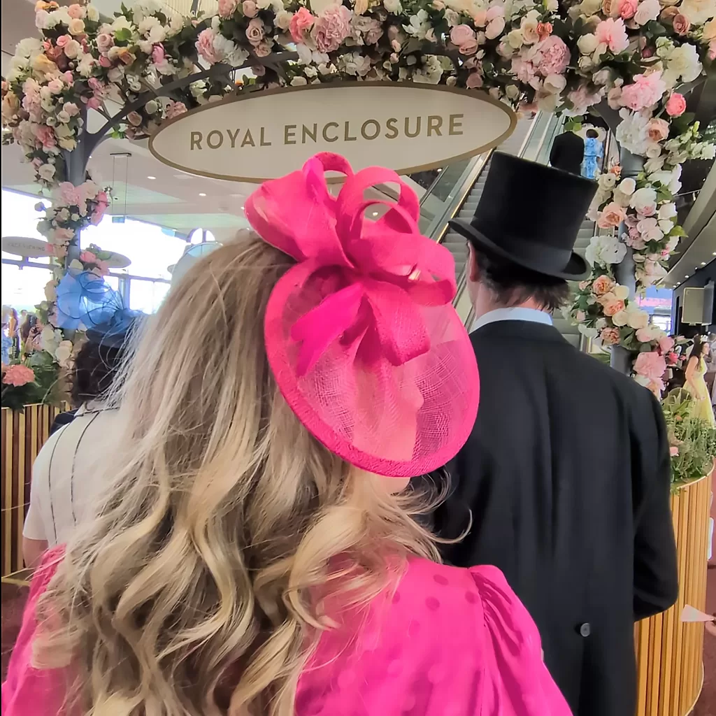 A woman in a bright pink fascinator and dress stands beside a man in a black top hat and suit, facing a floral arch with a sign that reads Royal Enclosure.