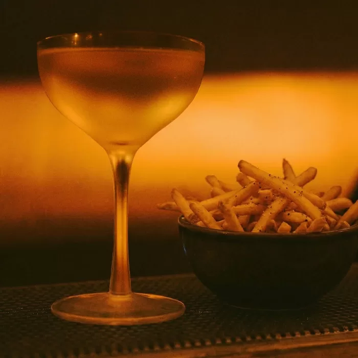 A cocktail glass filled with a golden drink sits beside a dark bowl of French fries on a dark surface, warmly lit by an amber light in the background.