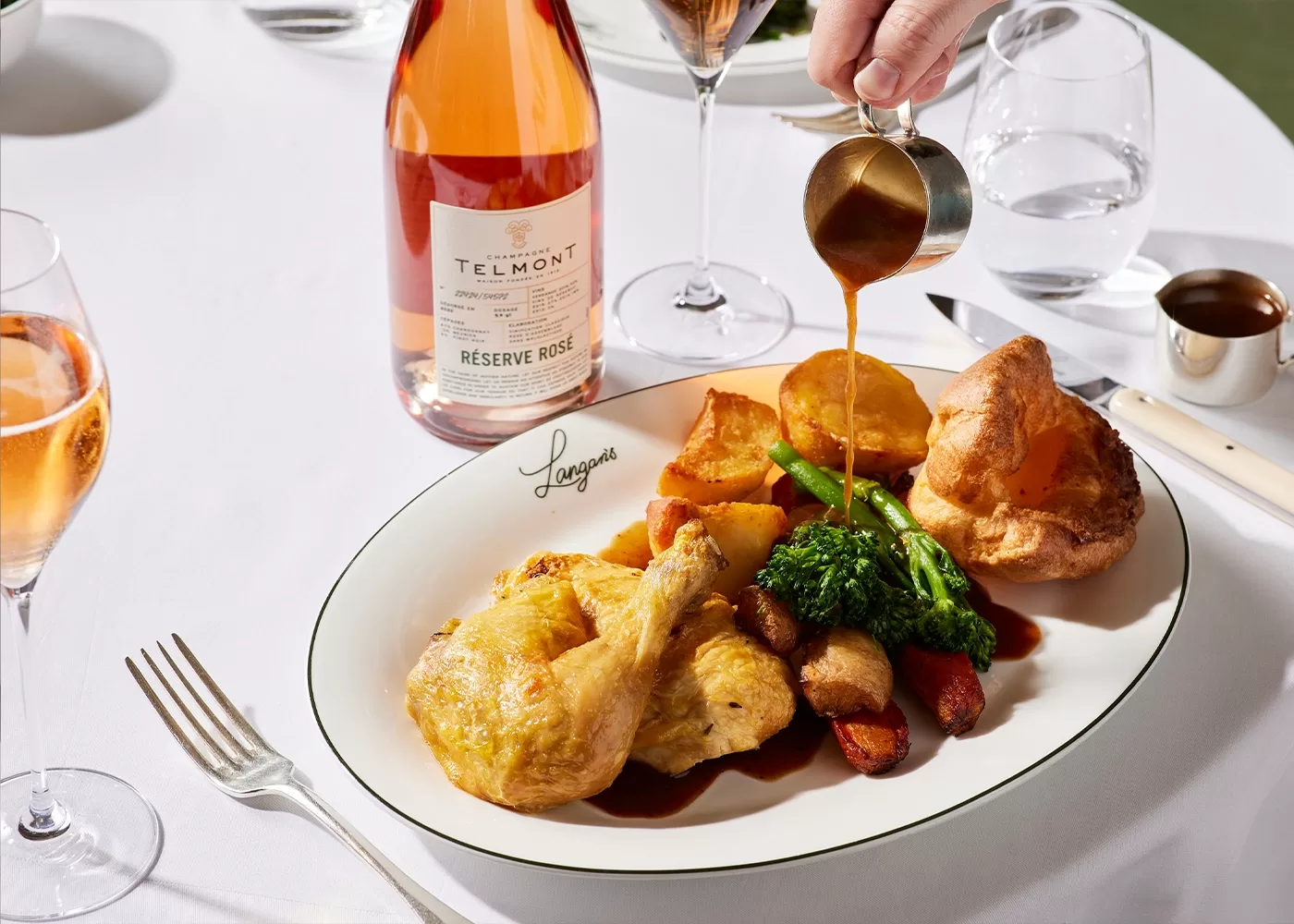 A plate of roast chicken, potatoes, vegetables, and Yorkshire pudding is topped with gravy. A hand pours gravy from a small jug. A glass and bottle of rosé wine, and glasses of water, are on a white-clothed table.