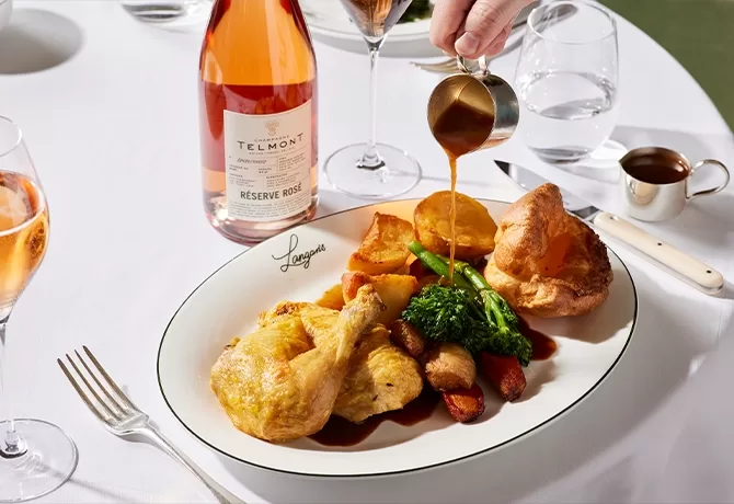 A roast chicken dinner with potatoes, carrots, broccoli, and Yorkshire pudding is served on a plate. Gravy is being poured over the meal. A bottle of rosé wine and glasses sit on the white-clothed table.