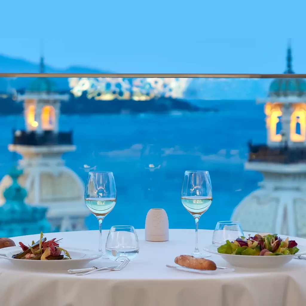 A table for two set with salad plates, bread, wine glasses, and water glasses overlooks a scenic view of domed buildings and a blue sea under a clear sky.