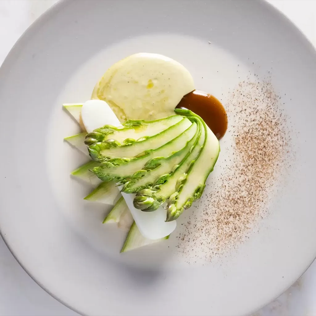 A gourmet dish on a white plate featuring thinly sliced asparagus, white sauce, brown sauce, and a dusting of spices, all artfully arranged with minimalist presentation.