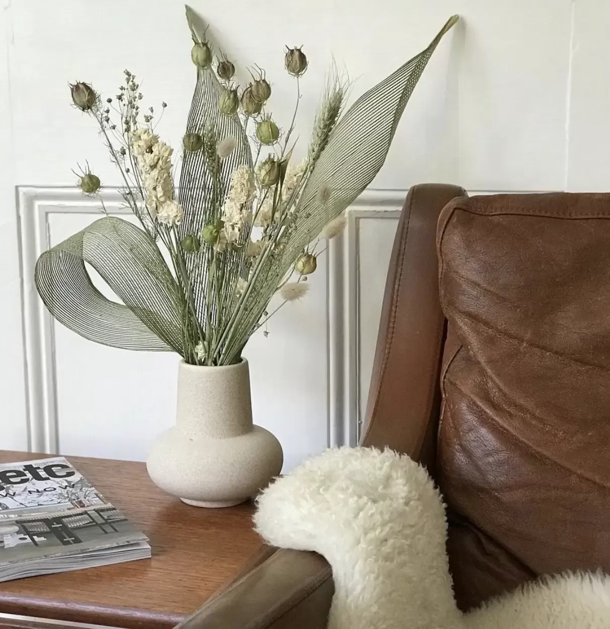 Elegant monochrome dried flowers in cream sculptural vase on a wooden bed side table next to brown leather sofa with fluffy blanket