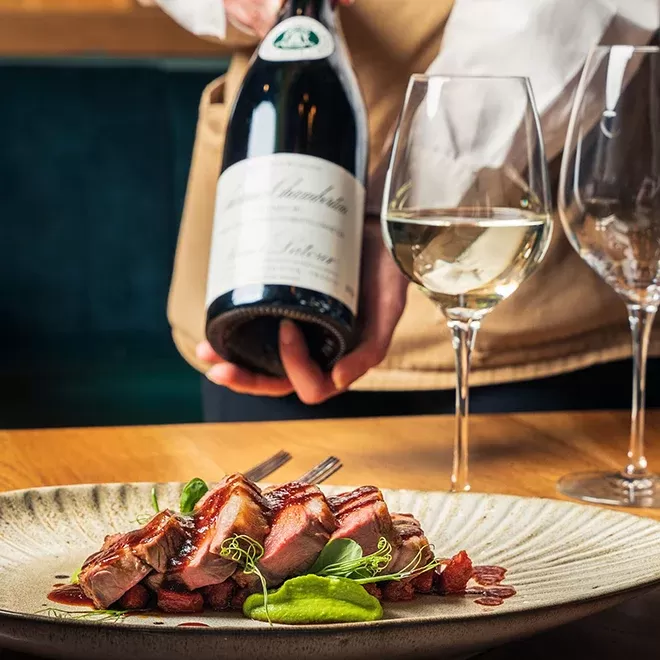 A person in a white shirt and apron holds a wine bottle next to a dining table. On the table, theres a plate of sliced meat with garnish and two wine glasses, one filled with white wine at Llanrech, Wales.