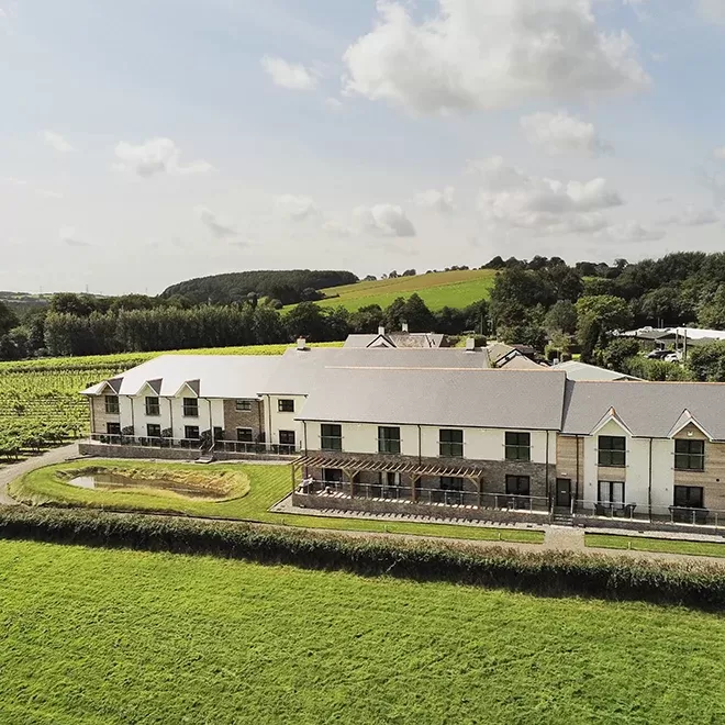 Aerial view of a large modern building surrounded by lush green fields and trees under a partly cloudy sky. The structure has a gray roof and white walls, with a small pond visible nearby. The landscape is picturesque and serene at Llanrech, Wales.