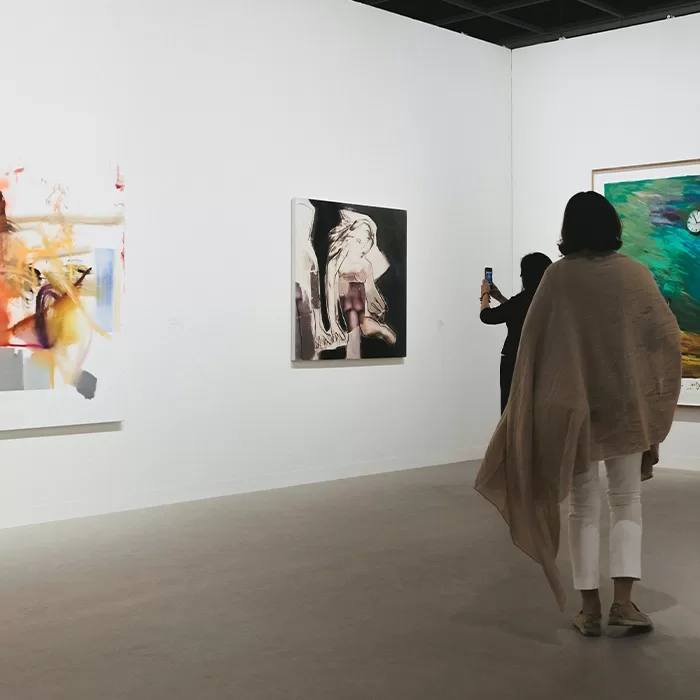 Two people view contemporary art on white gallery walls; one person takes a photo of a painting with a phone while another stands nearby, partially draped in a light shawl.