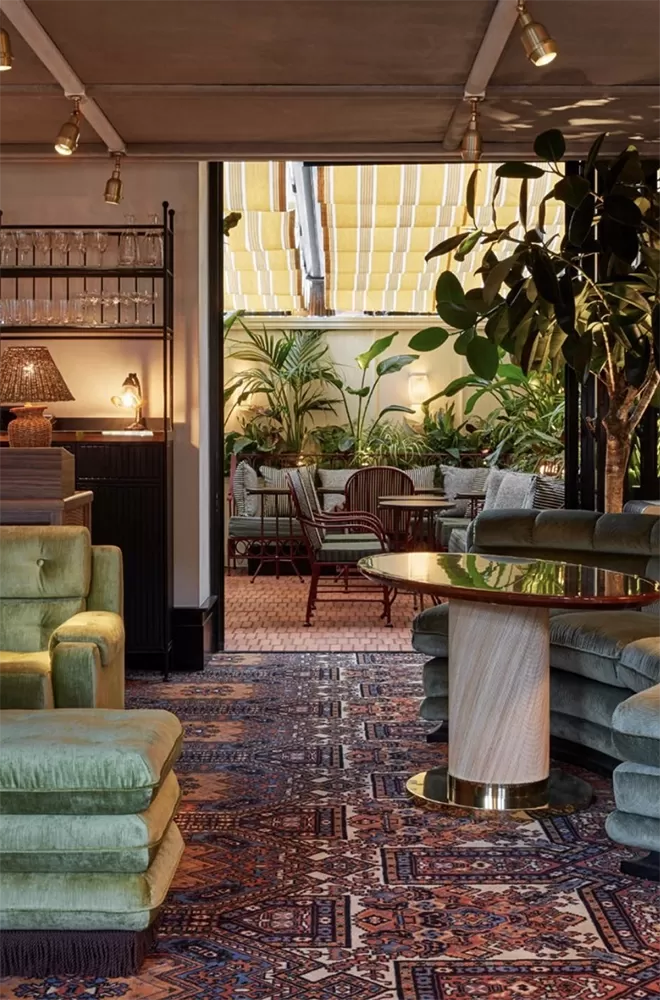 A chic indoor lounge with plush green sofas and patterned rugs opens to an outdoor seating area. The patio features red wicker chairs and lush plants under a striped awning, creating a cozy, tropical atmosphere at Louie, London.